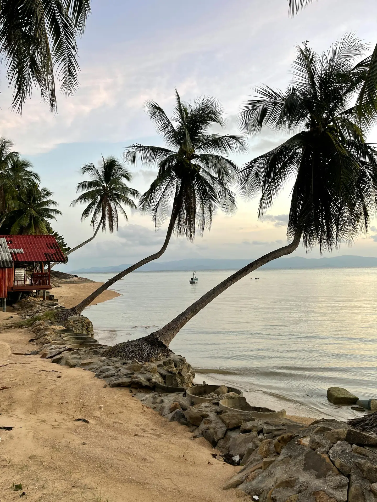 A quiet Thai beach with leaning palm trees at sunset, calm water reflecting the soft sky