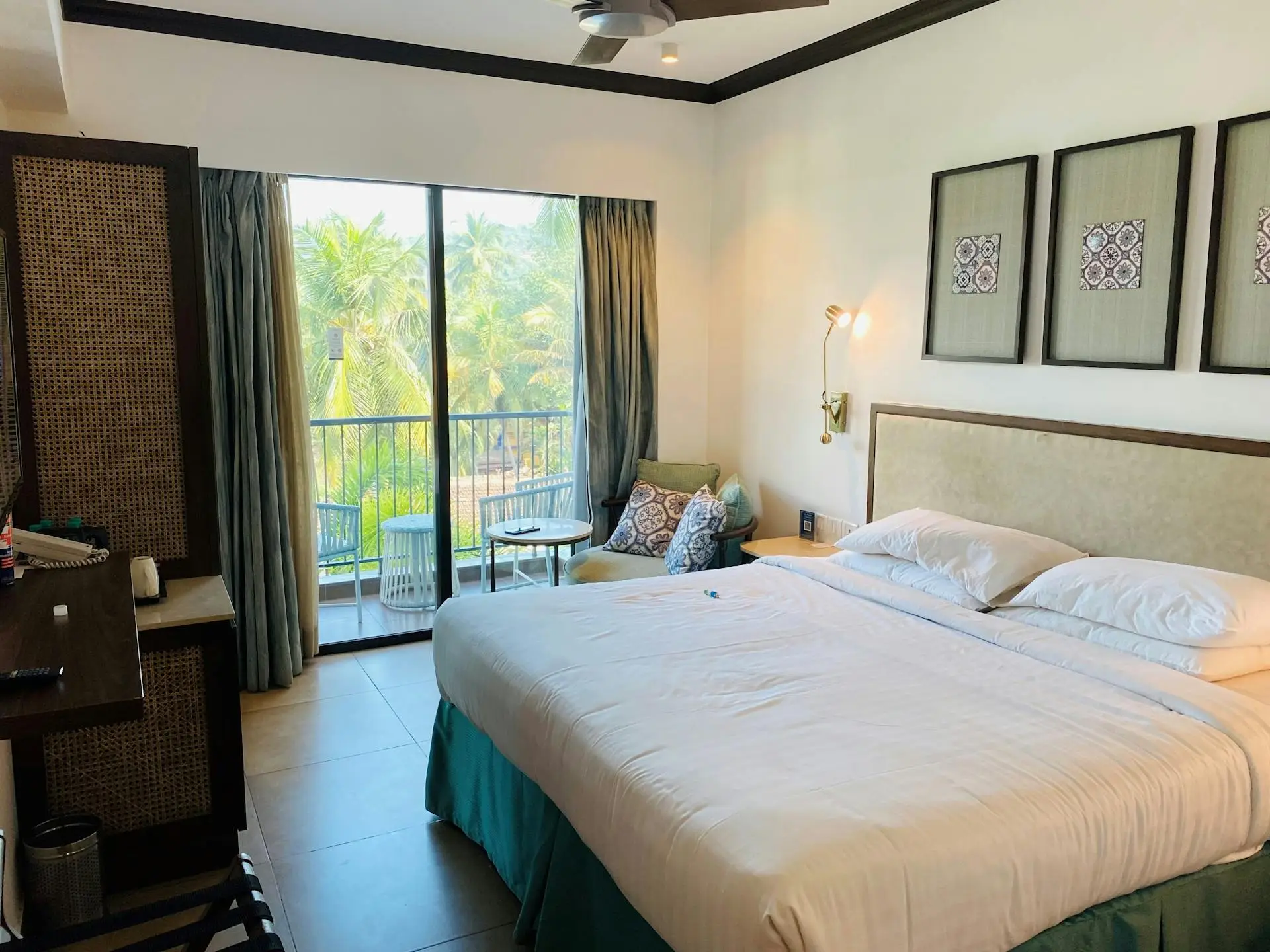 A bright, modern hotel room with white bedding, ceiling fan, and a balcony overlooking palm trees
